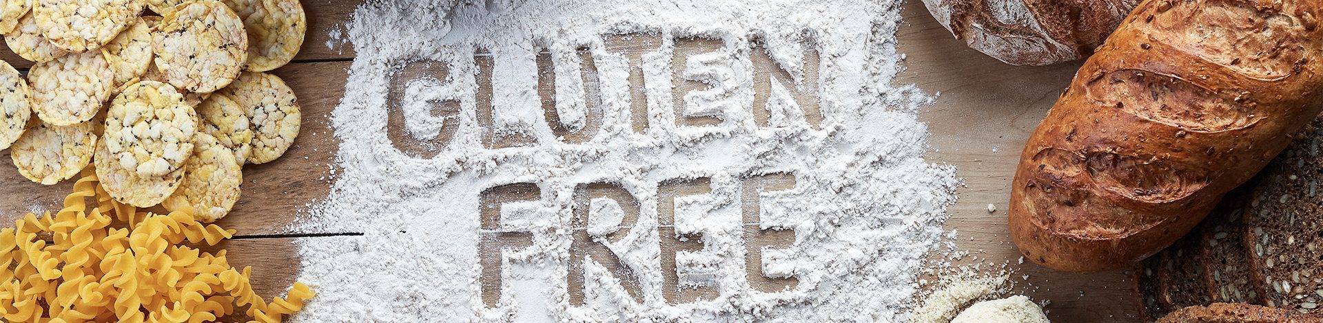 gluten-free bread, pasta and cakes on a messy table that reads 'gluten free' written in spilt flour