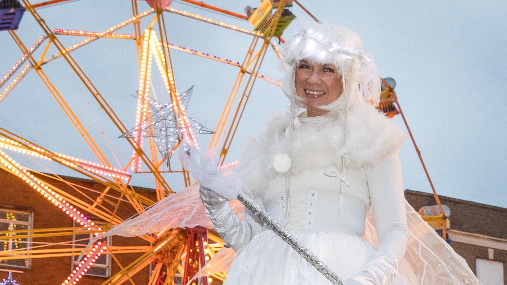 A lady dressed in white sparkly clothes and a white wig stands on stilts in front of the Ferris wheel
